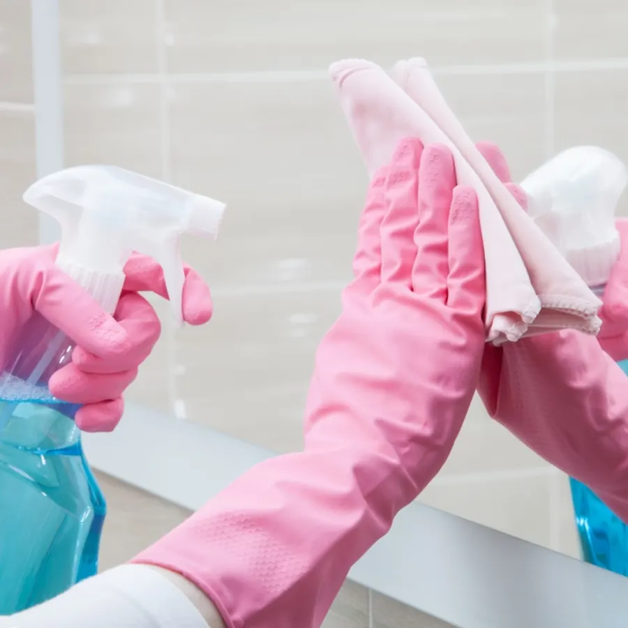 Pink gloved hands cleaning a bathroom mirror with a spray bottle and soft cloth