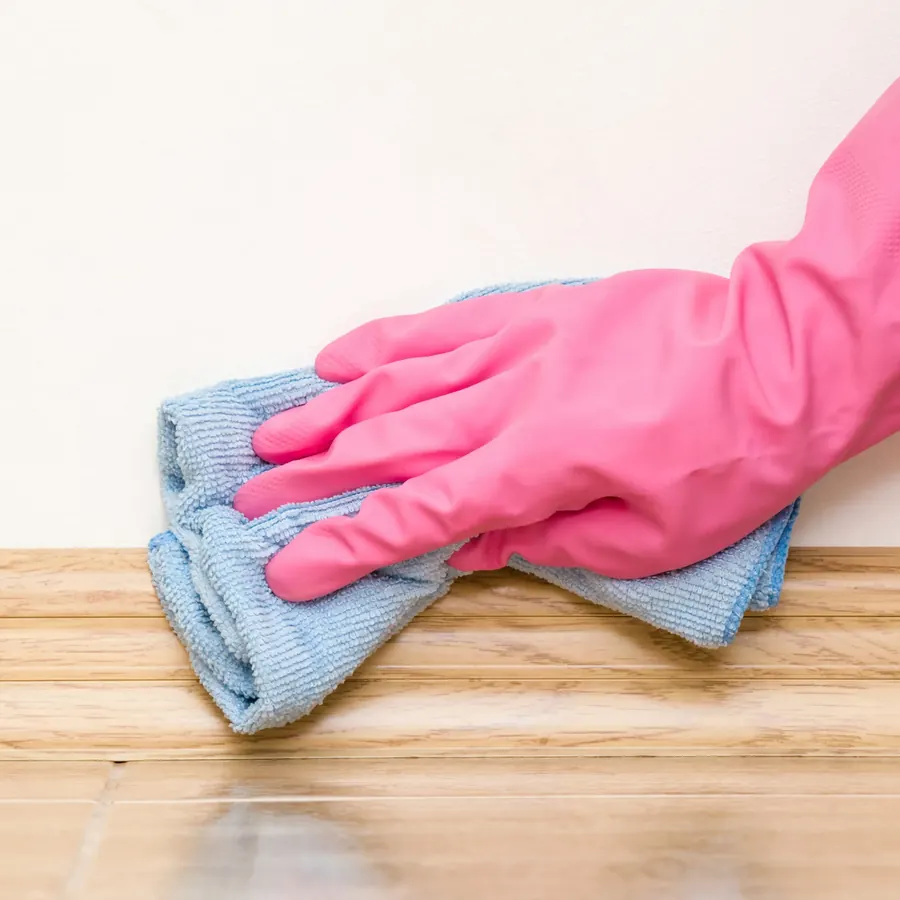 Gloved hand wiping a wood baseboard with a blue microfiber cloth