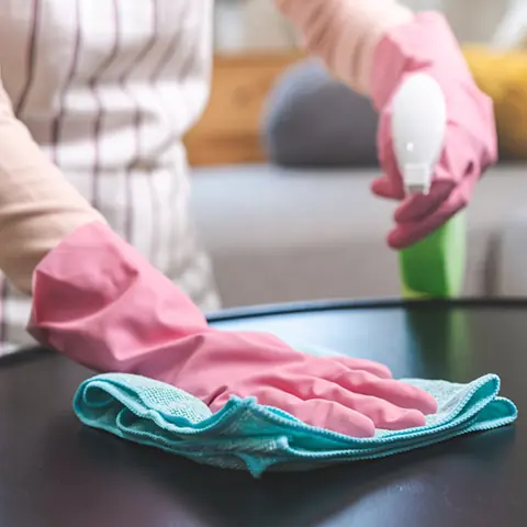 Pink gloved hand wiping a dark coffee table with a teal cloth in a living room