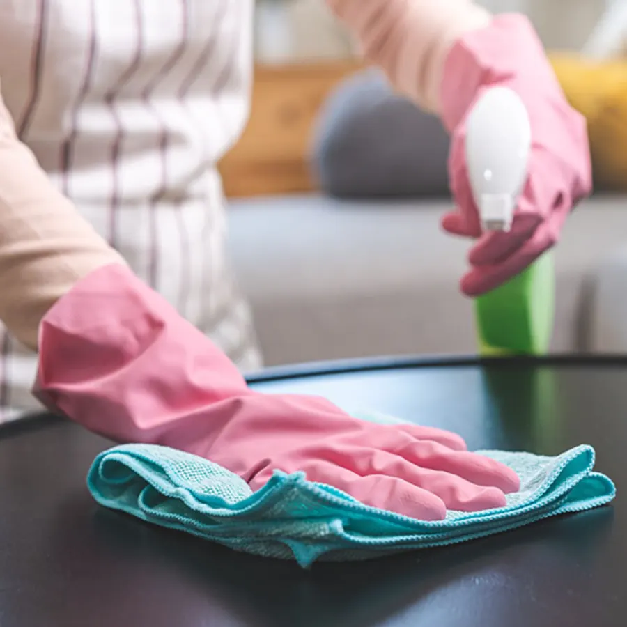 Pink gloved hand wiping a dark coffee table with a teal cloth in a living room
